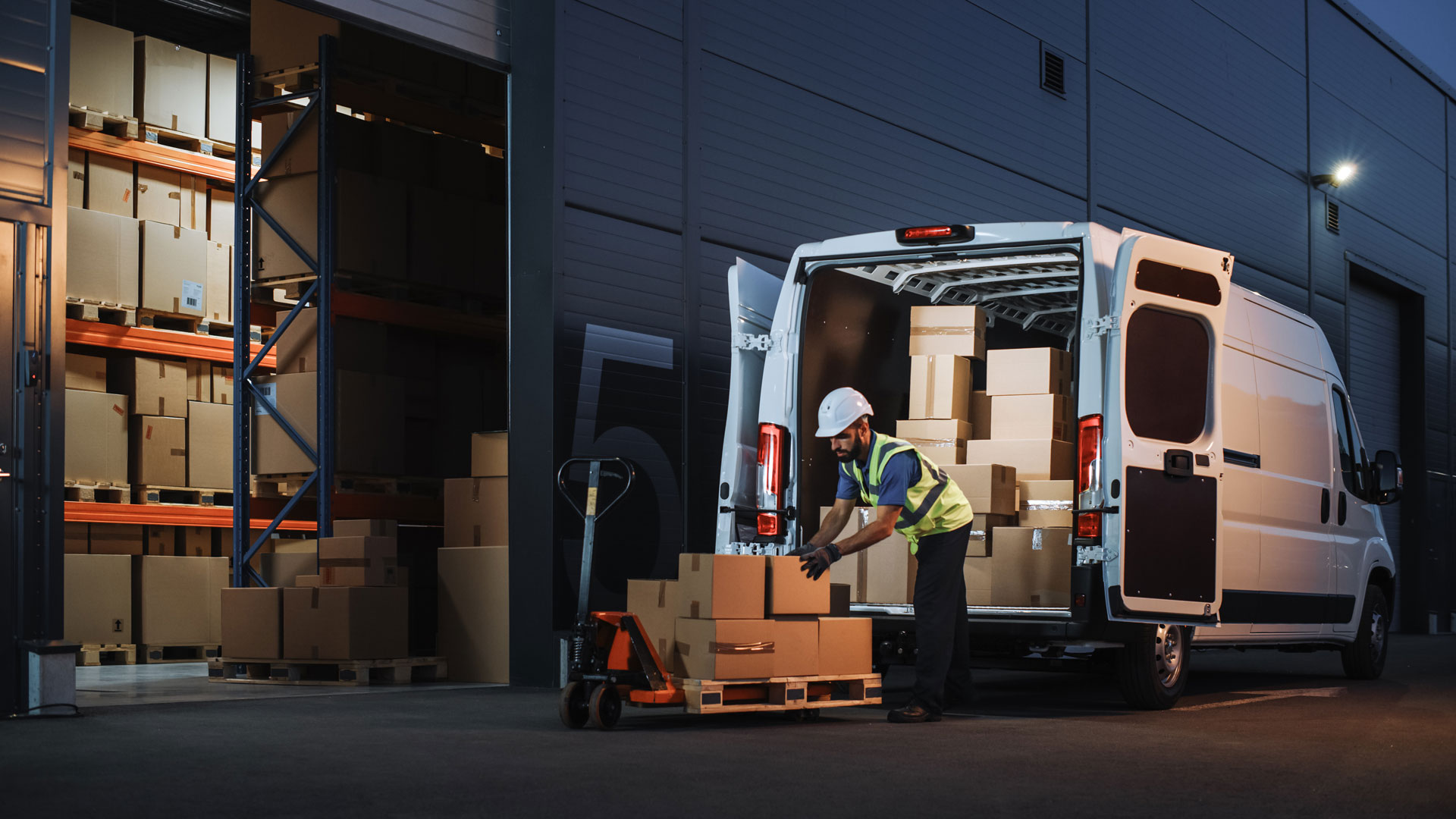 Outside of Logistics Distributions Warehouse Delivery Van: Worker Unloading Cardboard Boxes on Hand Truck, Online Orders, Purchases, E-Commerce Goods, Food, Medical Supply. Evening Shot 