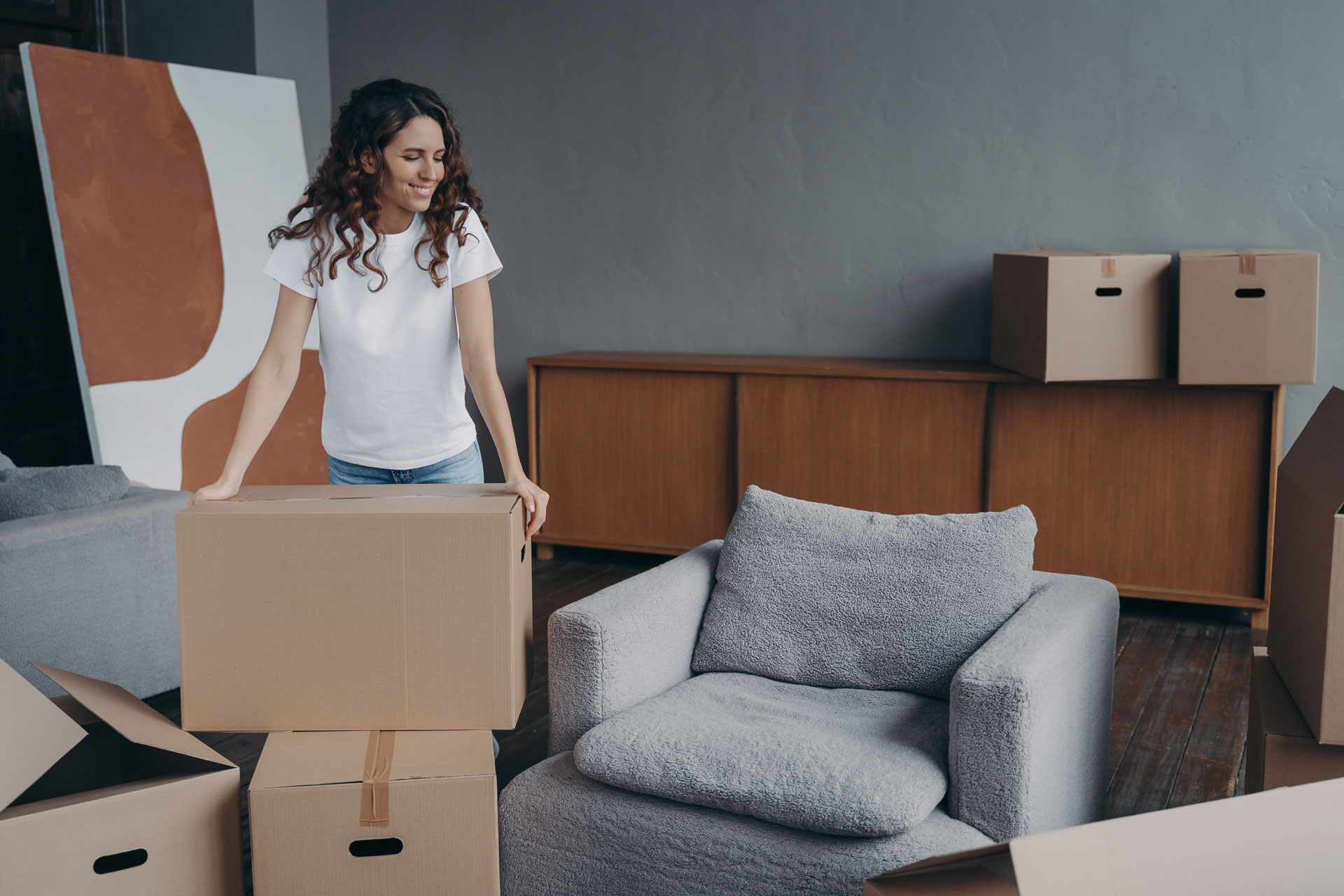 Smiling european woman is packing things. Young lady is unpacking cardboard boxes in new apartment.